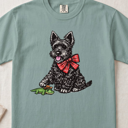 Sage green t-shirt featuring an illustration of a black terrier wearing a red plaid bow tie and touching a small toy alligator.