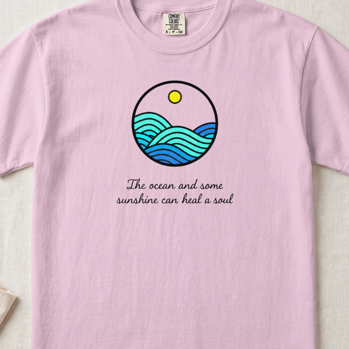 Pink t-shirt with a circular graphic of waves and sun, and text "The ocean and some sunshine can heal a soul".