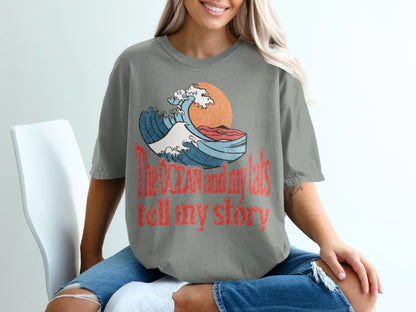 Gray t-shirt with graphic of crashing wave, sun, and text "The OCEAN and my tats tell my story".