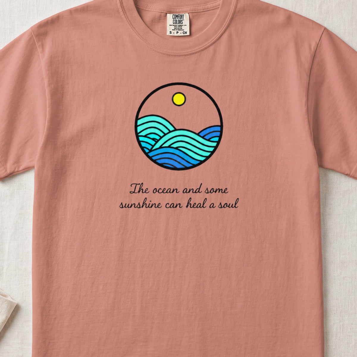 A terracotta t-shirt featuring a circular graphic of blue ocean waves and a yellow sun, with the text The ocean and some sunshine can heal a soul below.