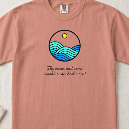 A terracotta t-shirt featuring a circular graphic of blue ocean waves and a yellow sun, with the text The ocean and some sunshine can heal a soul below.