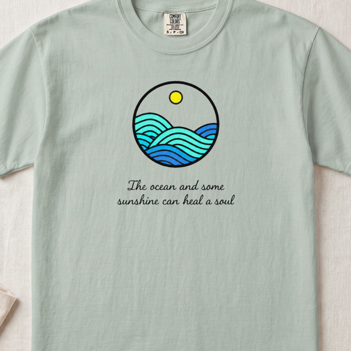 Light green t-shirt featuring a round ocean and sun graphic above the text The ocean and some sunshine can heal a soul.