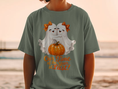 Two ghosts hug a pumpkin on a t-shirt with text "Our Time of the Year, at Last!".
