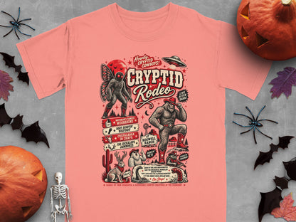Salmon pink t-shirt featuring a "Cryptid Rodeo" graphic with various mythical creatures and text.