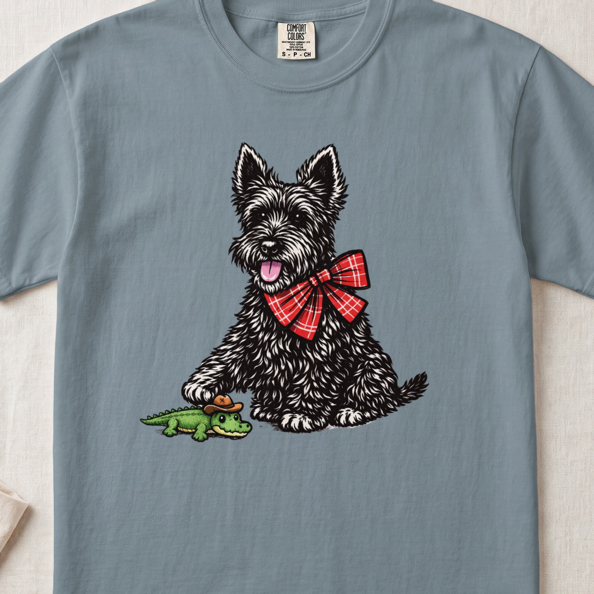 Blue t-shirt featuring an illustration of a black terrier wearing a red plaid bow tie, touching a small toy alligator.