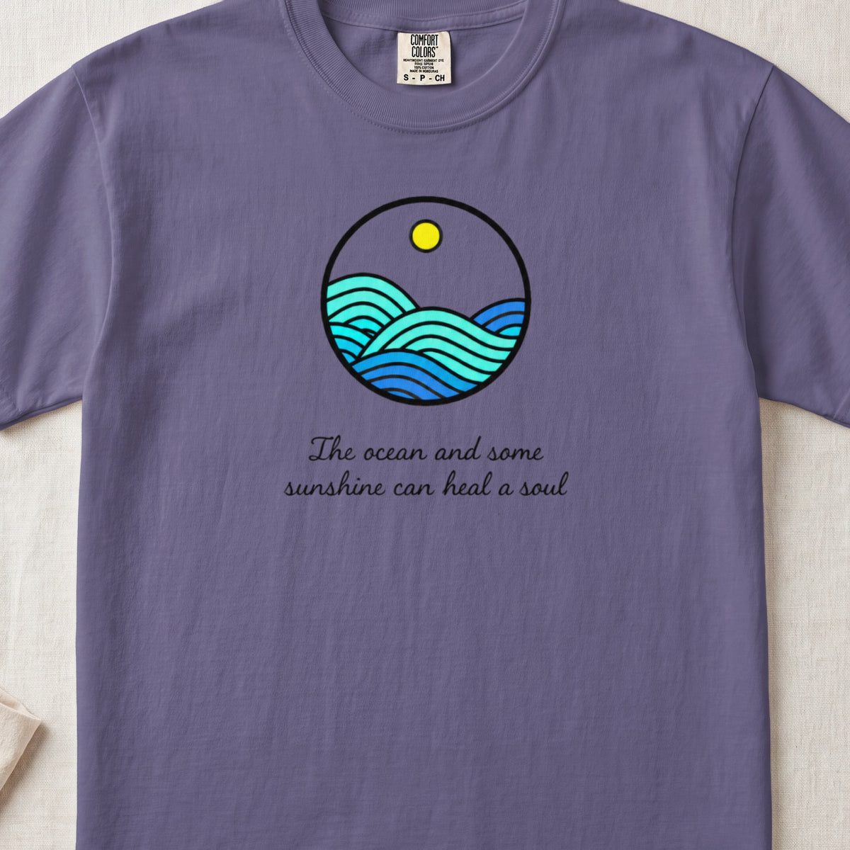 A purple t-shirt featuring a circular graphic of ocean waves and sunshine, with the text The ocean and some sunshine can heal a soul.