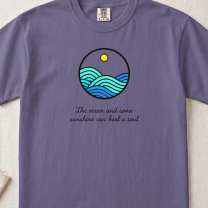 A purple t-shirt featuring a circular graphic of ocean waves and sunshine, with the text The ocean and some sunshine can heal a soul.