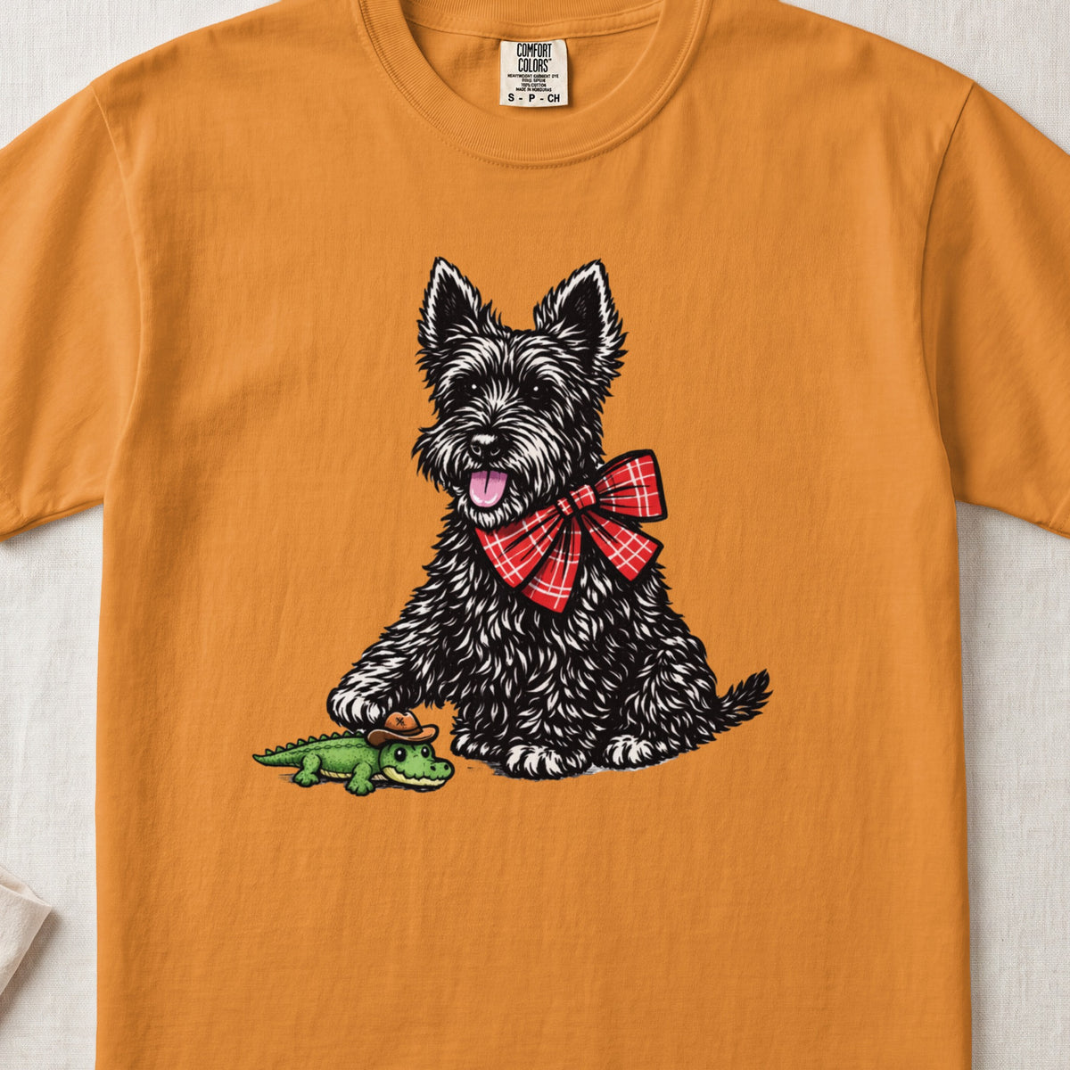 An orange crewneck t-shirt featuring an illustration of a black terrier wearing a red plaid bow, touching a small toy alligator with a cowboy hat.