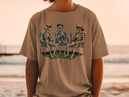 Three skeletons sit in lawn chairs holding drinks with a beach sunset in the background.