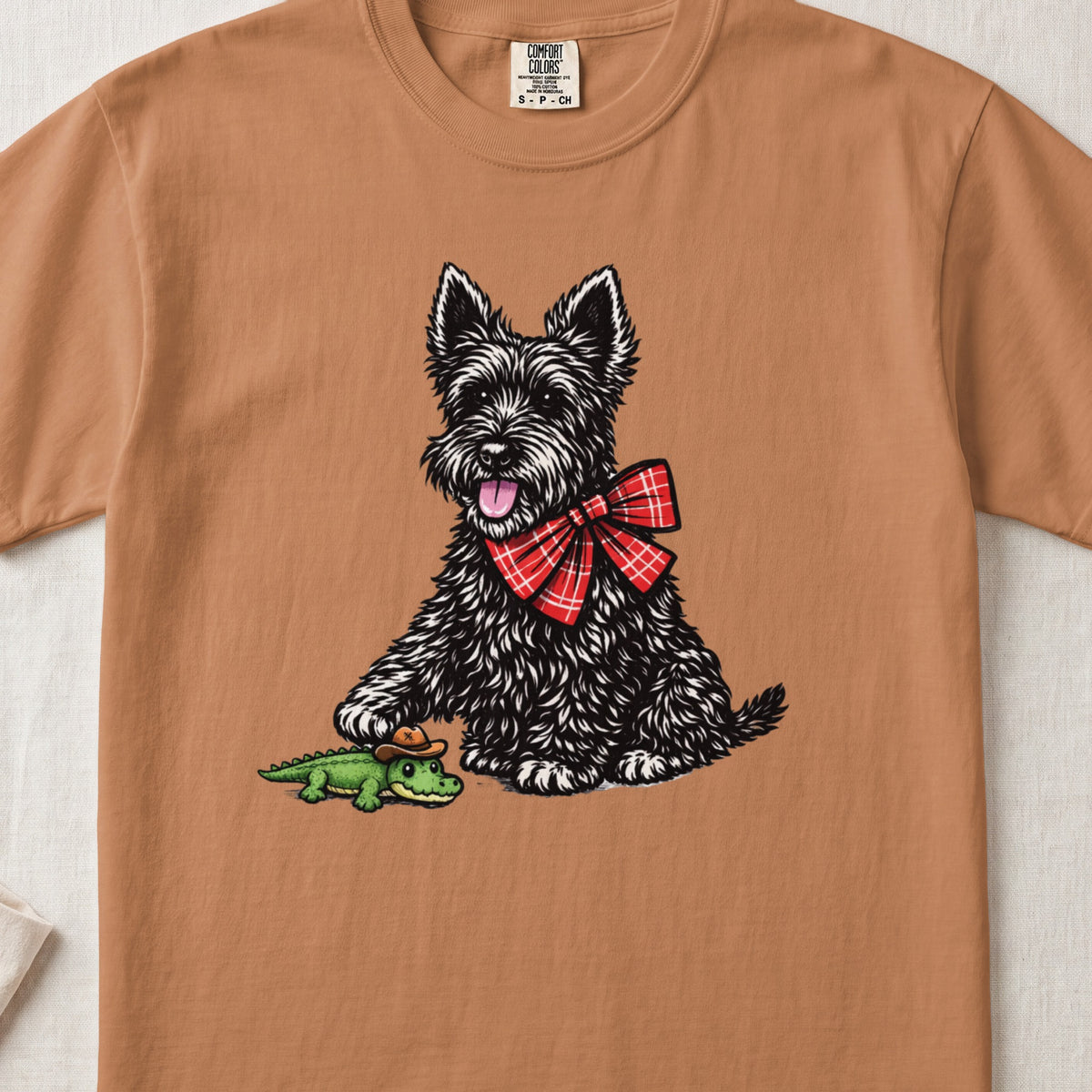 A brownish-orange t-shirt featuring an illustration of a black terrier wearing a red plaid bow, touching a small toy alligator.
