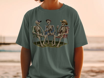 Three skeletons sit in lawn chairs on grass, holding drinks.