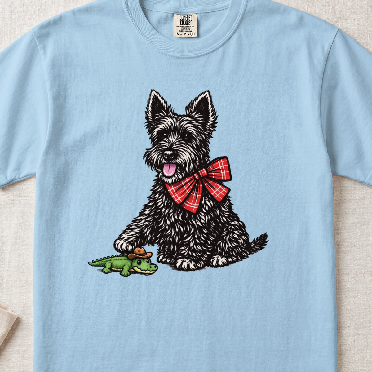 Light blue t-shirt featuring an illustration of a black terrier wearing a red plaid bow tie, stepping on a toy alligator.