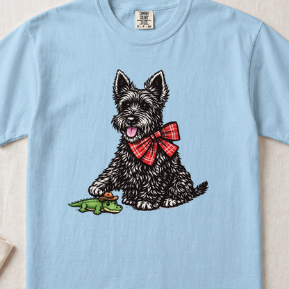 Light blue t-shirt featuring an illustration of a black terrier wearing a red plaid bow tie, stepping on a toy alligator.