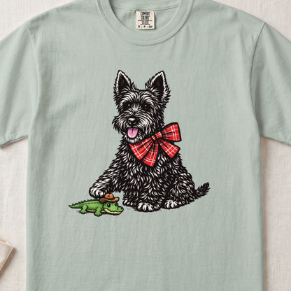 A sage green t-shirt featuring an illustration of a black Scottish Terrier wearing a red plaid bow, touching a toy alligator.