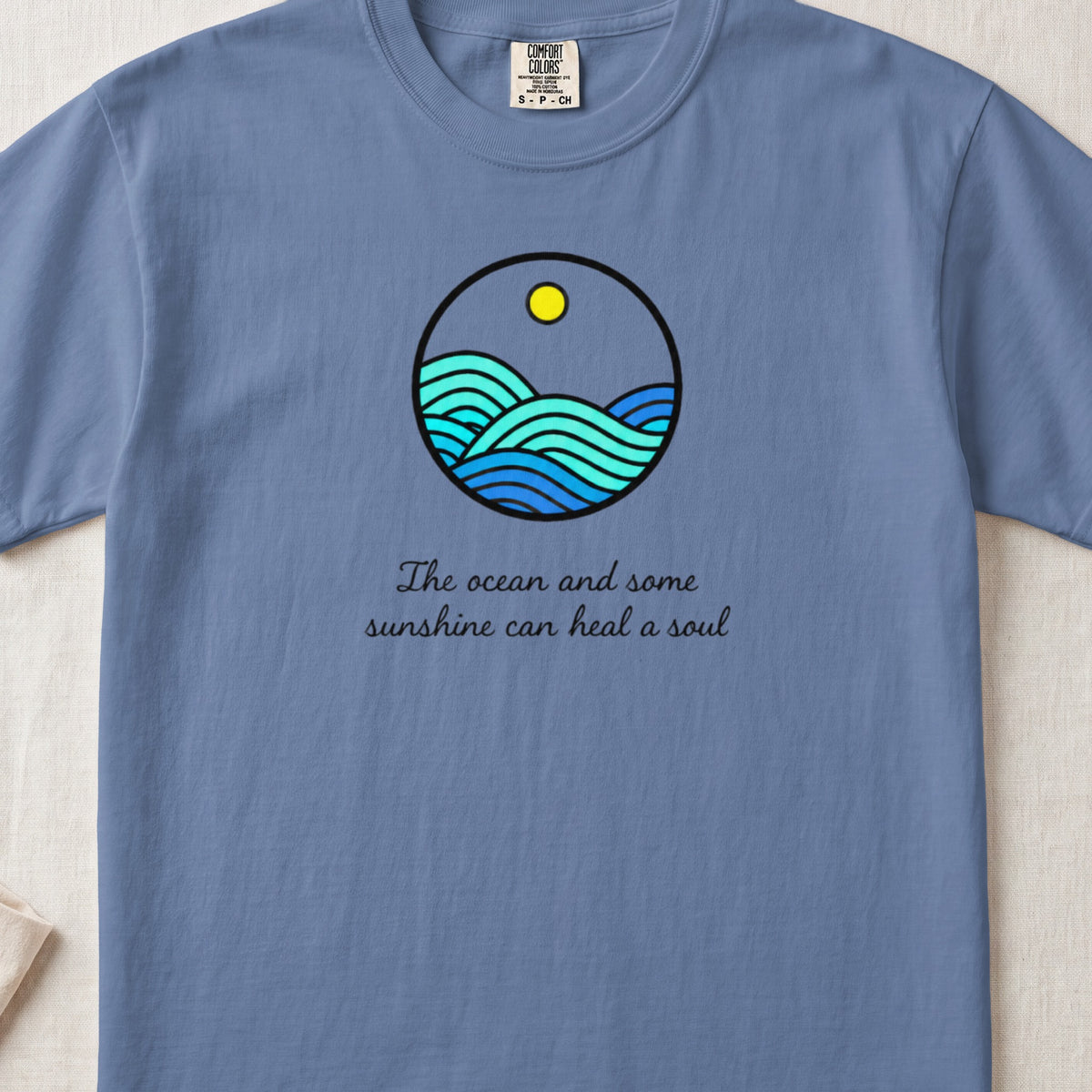 A blue crewneck t-shirt featuring a circular ocean graphic and the text The ocean and some sunshine can heal a soul.