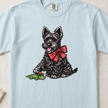 A light blue t-shirt featuring an illustration of a black terrier wearing a red plaid bow tie, playing with a small toy alligator wearing a cowboy hat.