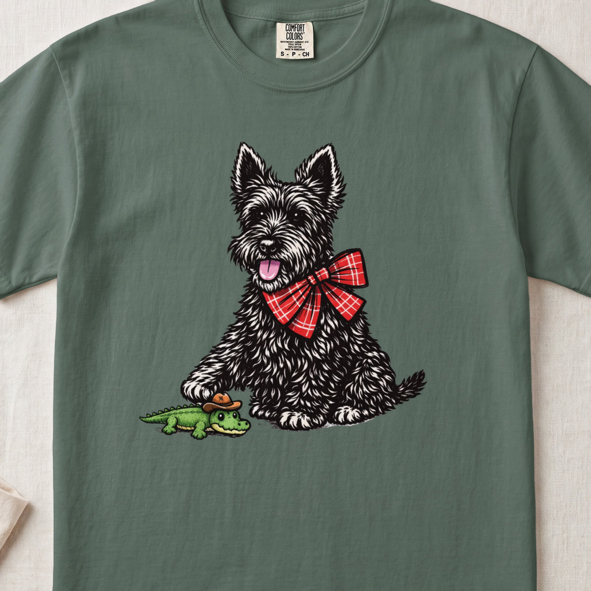 A green crewneck t-shirt featuring an illustration of a black terrier wearing a red plaid bow, touching a small alligator toy.