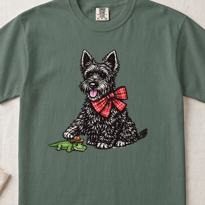 A green crewneck t-shirt featuring an illustration of a black terrier wearing a red plaid bow, touching a small alligator toy.