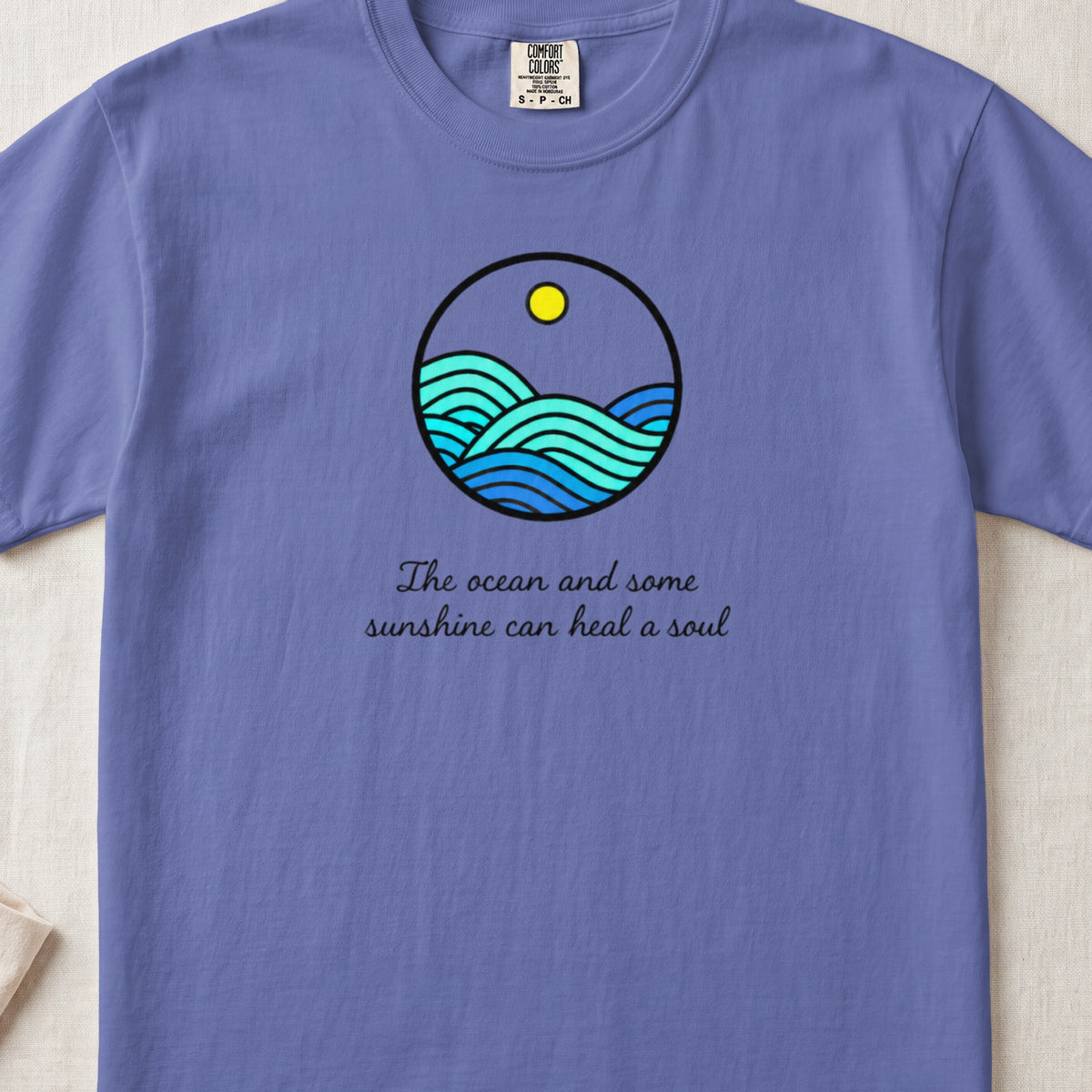 A periwinkle t-shirt features a circular ocean graphic with a yellow sun and text reading The ocean and some sunshine can heal a soul.