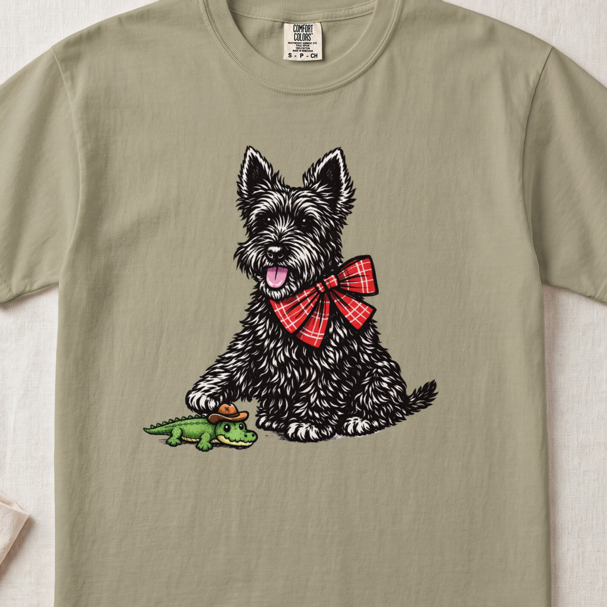 Olive green t-shirt featuring a graphic of a black terrier wearing a red plaid bow tie and touching a small alligator toy.