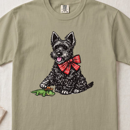 Olive green t-shirt featuring a graphic of a black terrier wearing a red plaid bow tie and touching a small alligator toy.