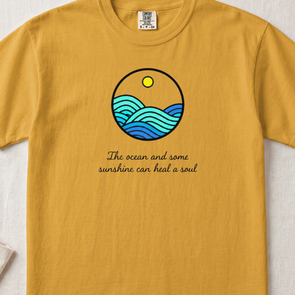A mustard yellow t-shirt featuring a circular ocean and sun graphic with cursive text underneath.