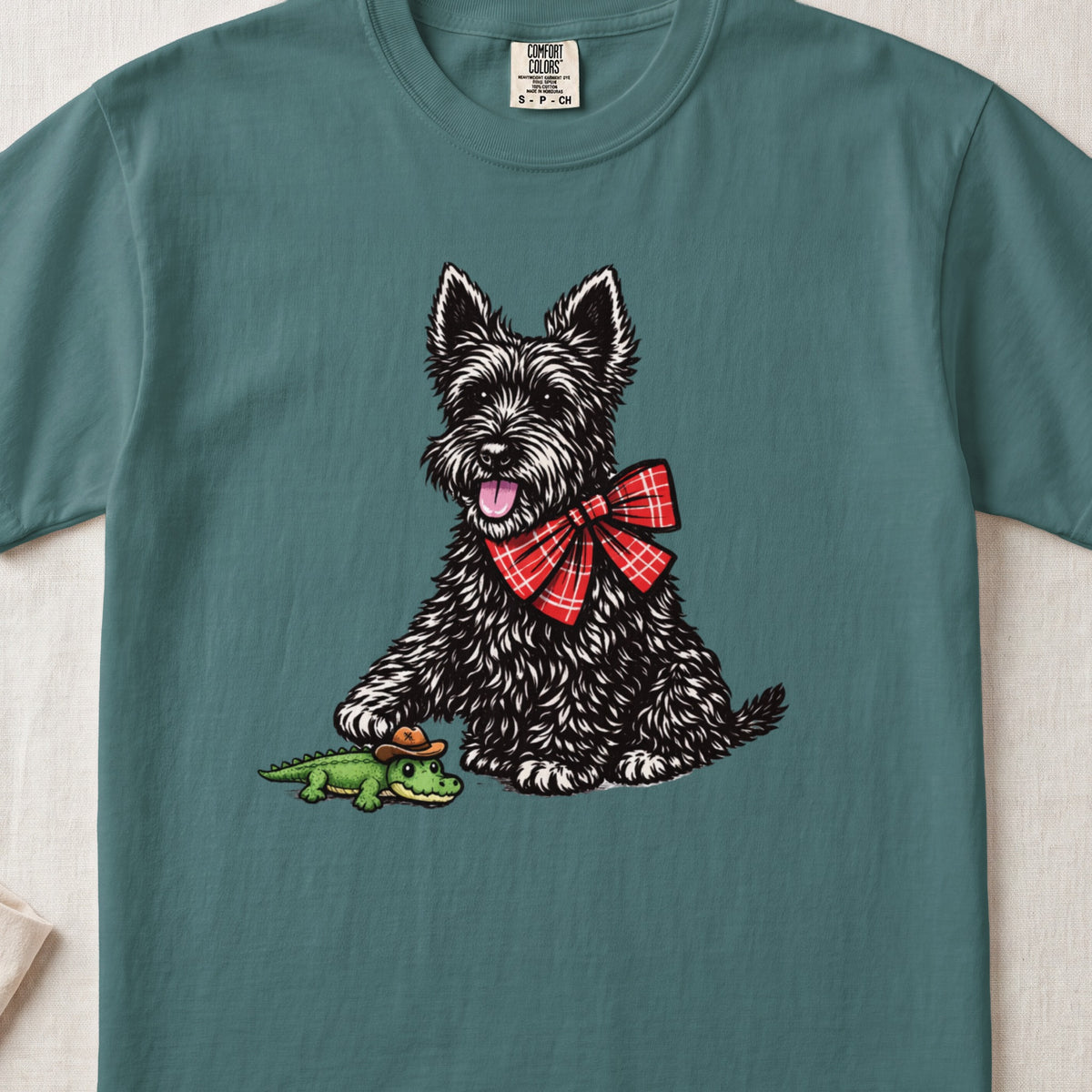 A teal t-shirt features an illustration of a black terrier wearing a red plaid bow, touching a tiny alligator wearing a cowboy hat.