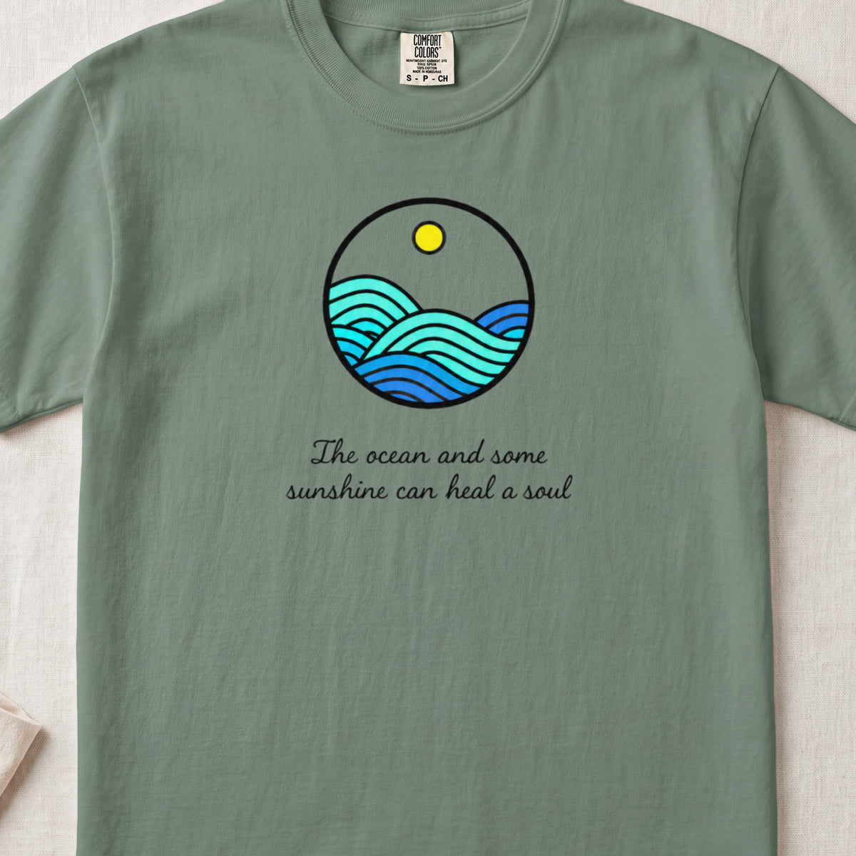 A sage green t-shirt featuring a circular ocean graphic and the text The ocean and some sunshine can heal a soul.