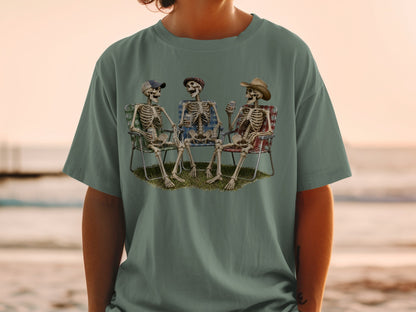 Three skeletons sitting in camping chairs on grass, drinking beverages.