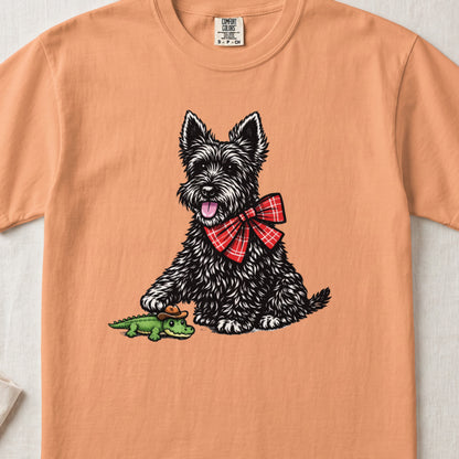 Peach-colored t-shirt featuring a black Scottish Terrier wearing a red plaid bow tie and touching a toy alligator.