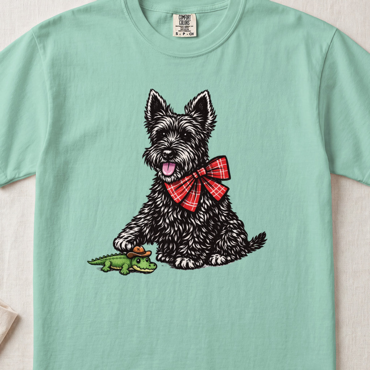 A light green t-shirt featuring an illustration of a black terrier wearing a red plaid bow, touching a toy alligator.