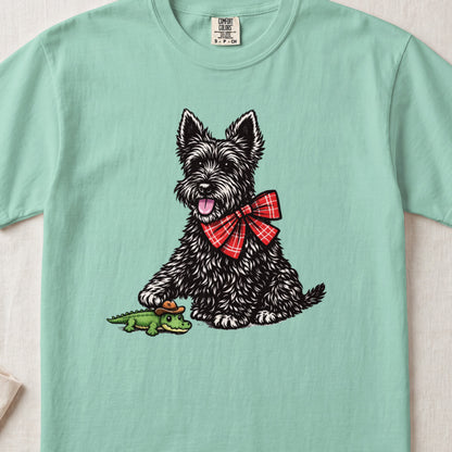 A light green t-shirt featuring an illustration of a black terrier wearing a red plaid bow, touching a toy alligator.