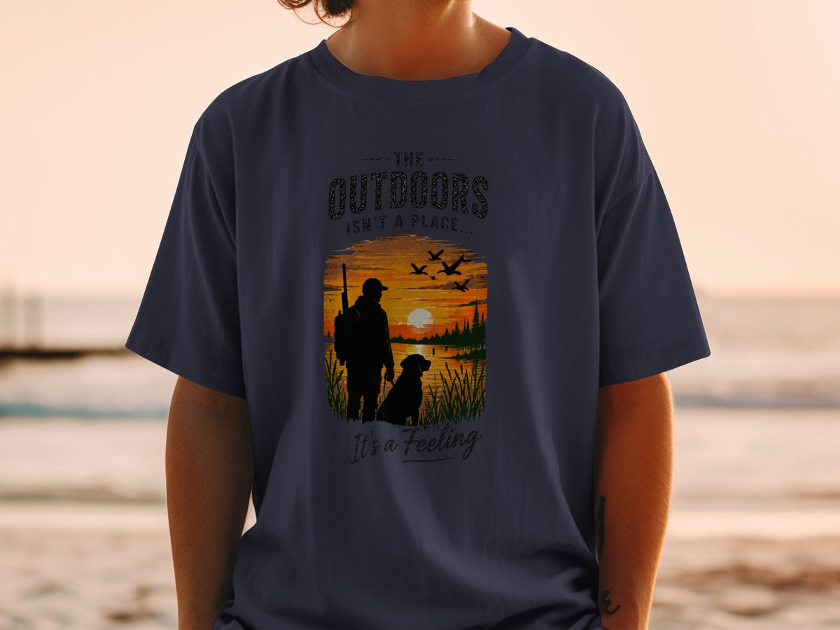 Navy t-shirt with graphic of hunter, dog, and geese at sunset, text reads "The Outdoors Isn't A Place... It's a Feeling".