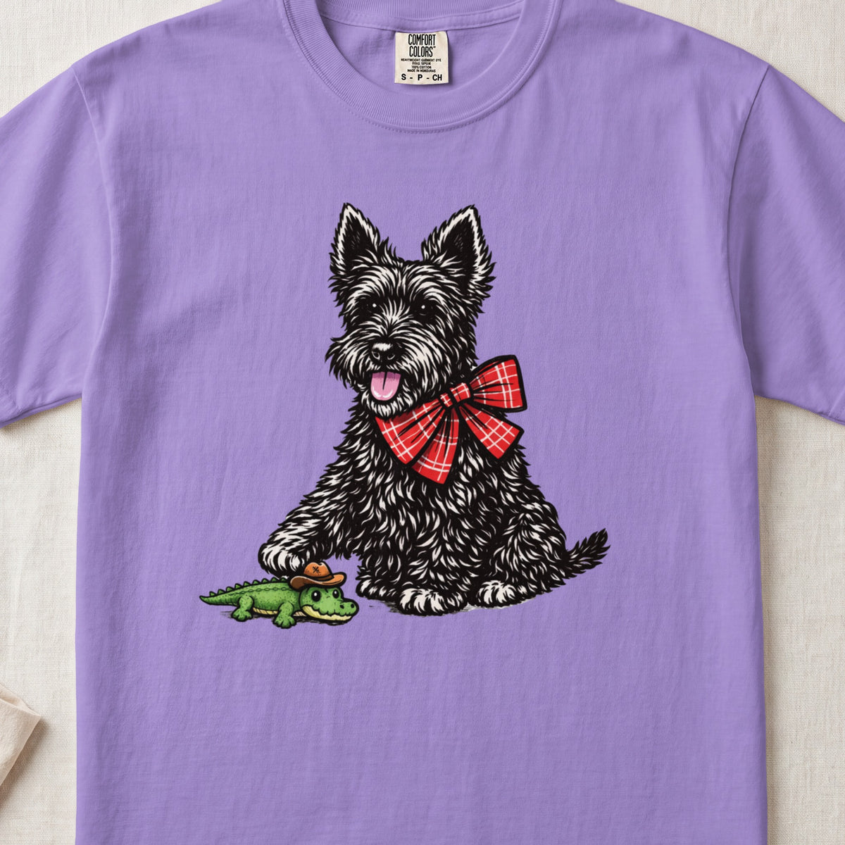 Light purple t-shirt featuring a black Scottish Terrier wearing a red plaid bow tie and touching a toy alligator.