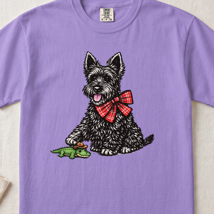 Light purple t-shirt featuring a black Scottish Terrier wearing a red plaid bow tie and touching a toy alligator.