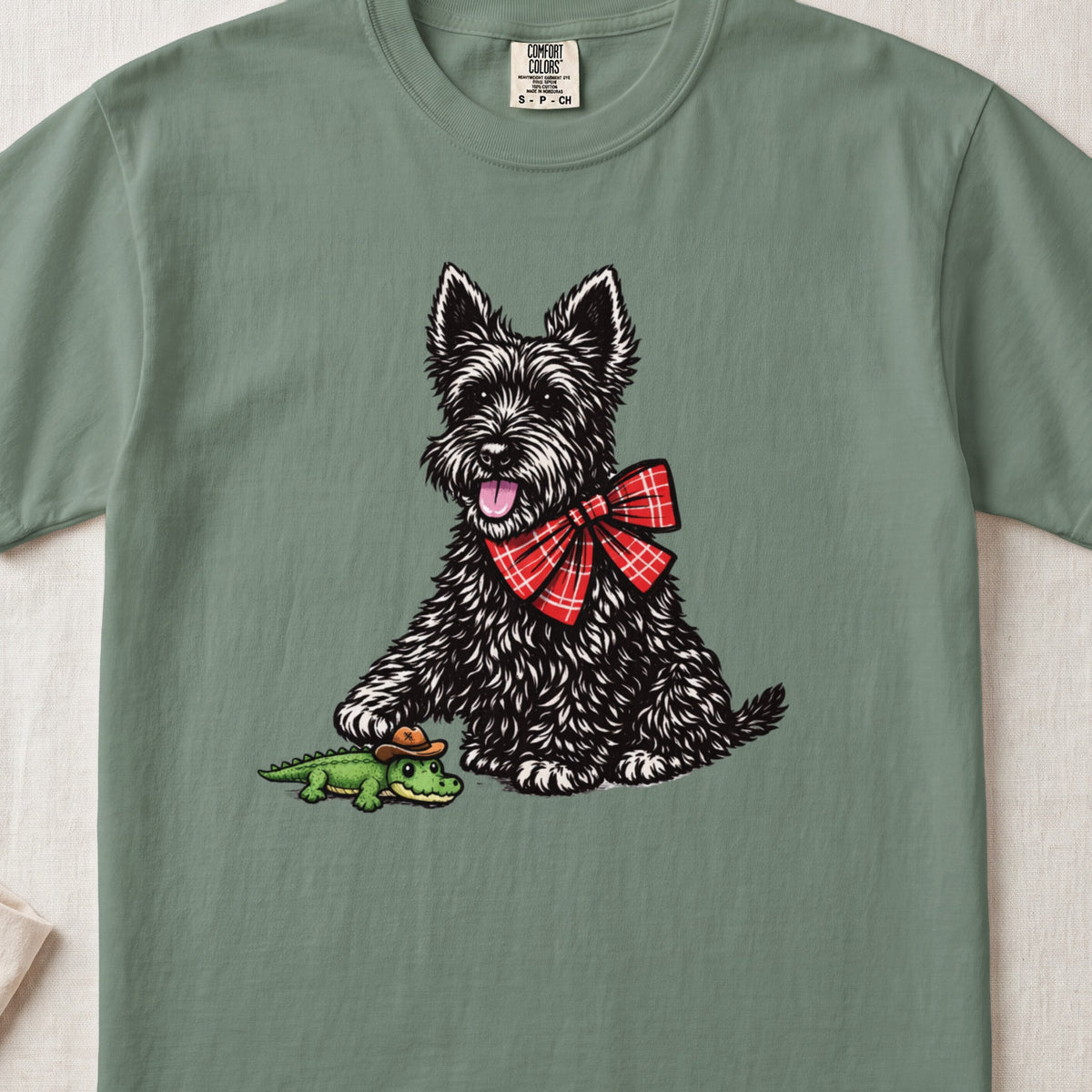 A sage green t-shirt featuring an illustration of a black Scottish Terrier wearing a red plaid bow, stepping on a toy alligator.