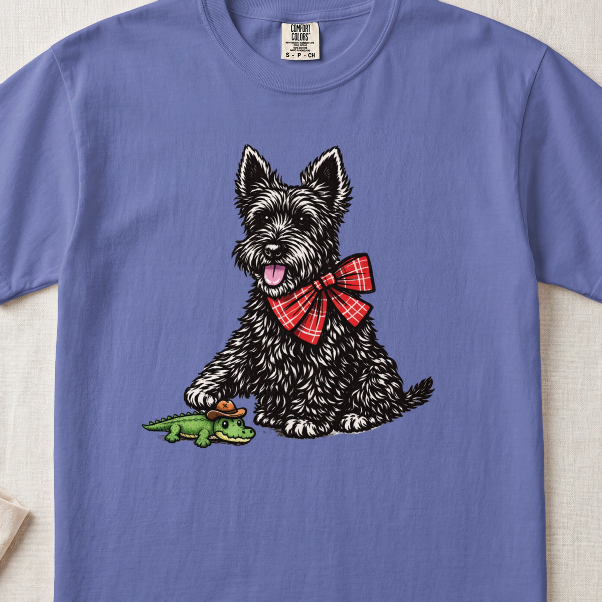 Periwinkle t-shirt featuring an illustration of a black terrier dog wearing a red plaid bow, holding a small toy alligator.