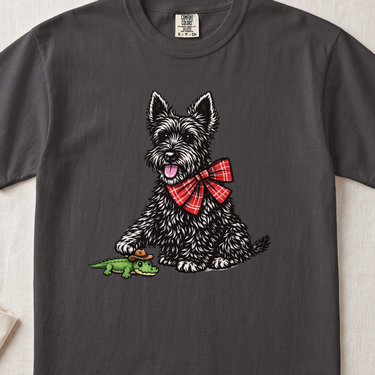 A dark gray t-shirt featuring a graphic of a black terrier wearing a red plaid bow, touching a toy alligator.