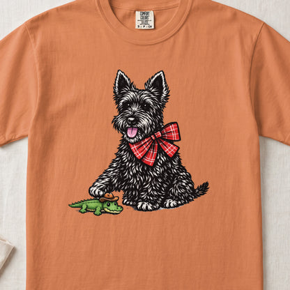 An orange t-shirt featuring a graphic of a black terrier wearing a red plaid bow, placing its paw on a small green alligator wearing a cowboy hat.