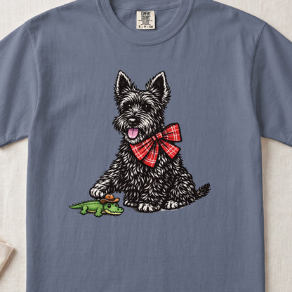 A grey t-shirt featuring an illustration of a black Scottish Terrier wearing a red plaid bow tie, touching a small green alligator toy wearing a cowboy hat.
