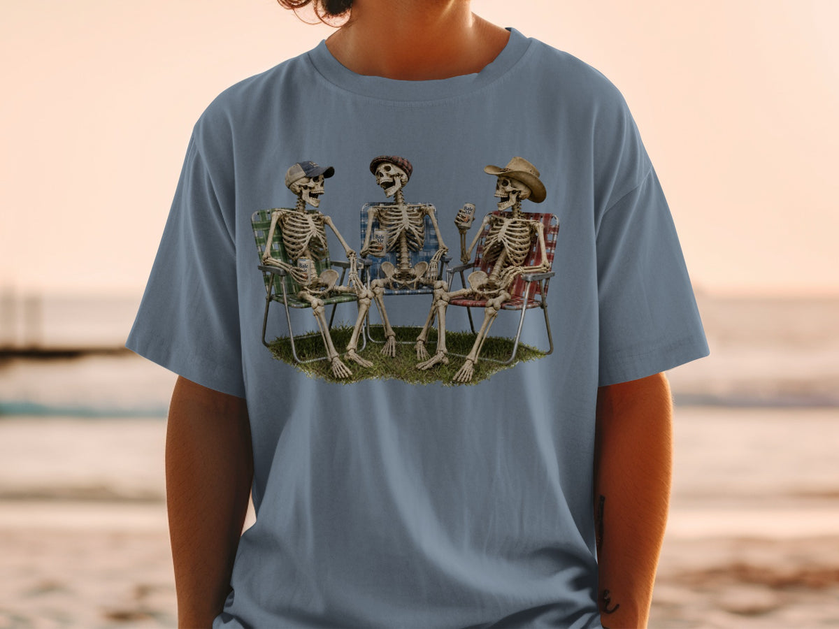Three skeletons in chairs on grass with drinks and hats, viewed on a t-shirt.