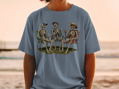 Three skeletons in chairs on grass with drinks and hats, viewed on a t-shirt.