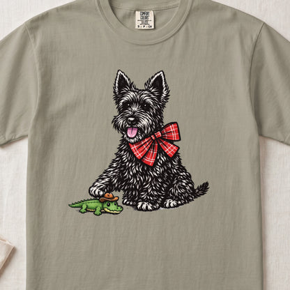 An olive green crewneck t-shirt featuring an illustration of a black Scottish Terrier wearing a red plaid bow tie, touching a small toy alligator with a cowboy hat.