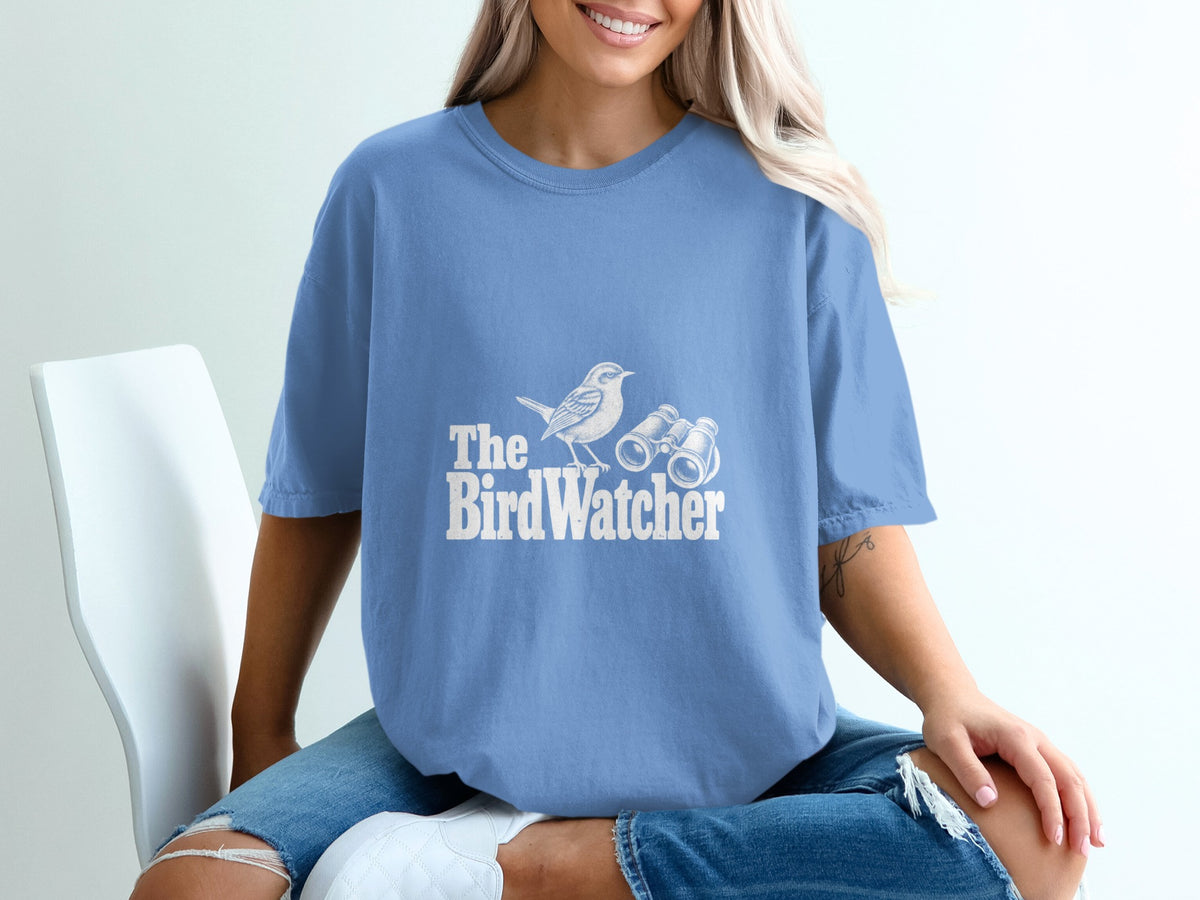 Blue t-shirt with white text and illustration: The BirdWatcher, a bird and binoculars.