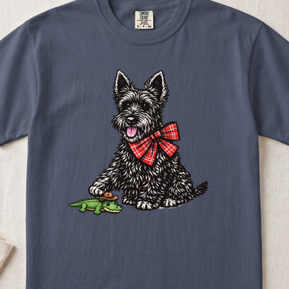 A slate blue t-shirt printed with a graphic of a black terrier wearing a red plaid bow, touching a small toy alligator wearing a cowboy hat.