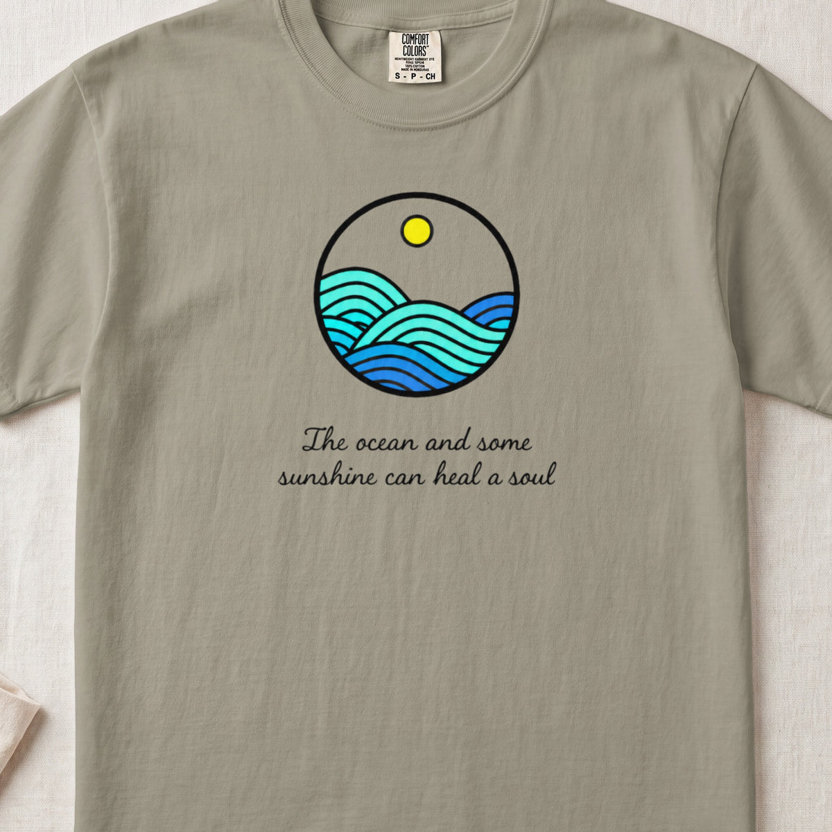 Sage green t-shirt featuring a circular ocean graphic with a sun and the text The ocean and some sunshine can heal a soul.