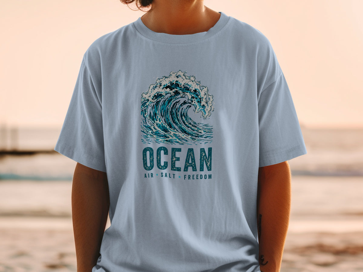 Light blue t-shirt with a crashing wave graphic and text reading "OCEAN AIR SALT FREEDOM".