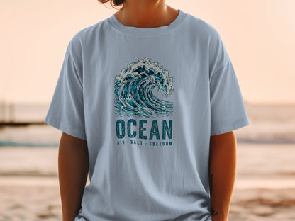 Light blue t-shirt with a crashing wave graphic and text reading "OCEAN AIR SALT FREEDOM".