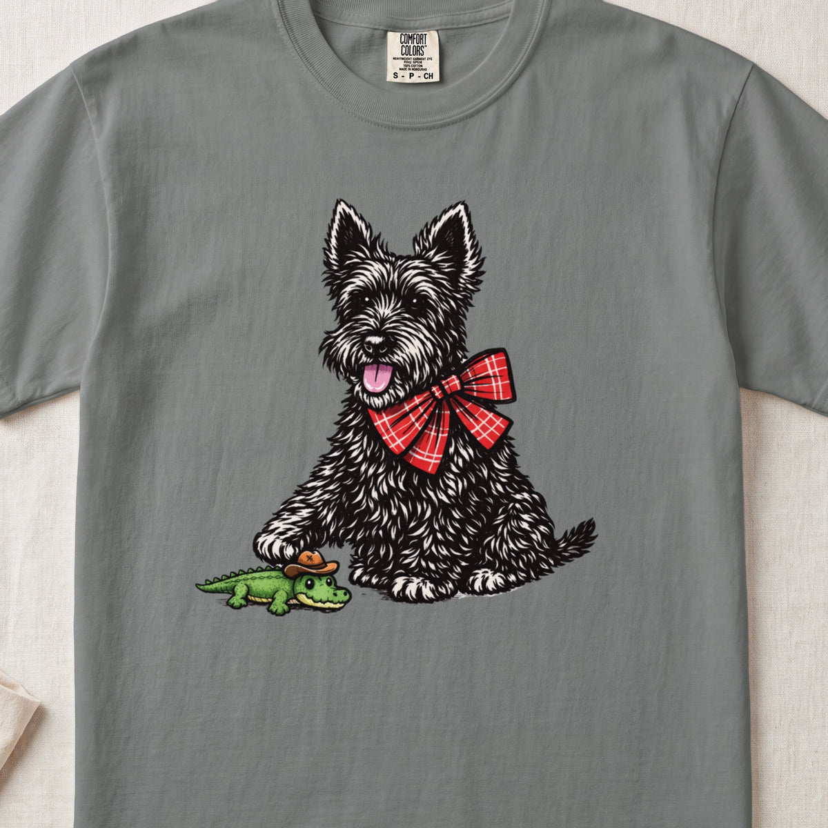 Grey t-shirt featuring an illustration of a black terrier wearing a red plaid bow tie and touching a toy alligator.
