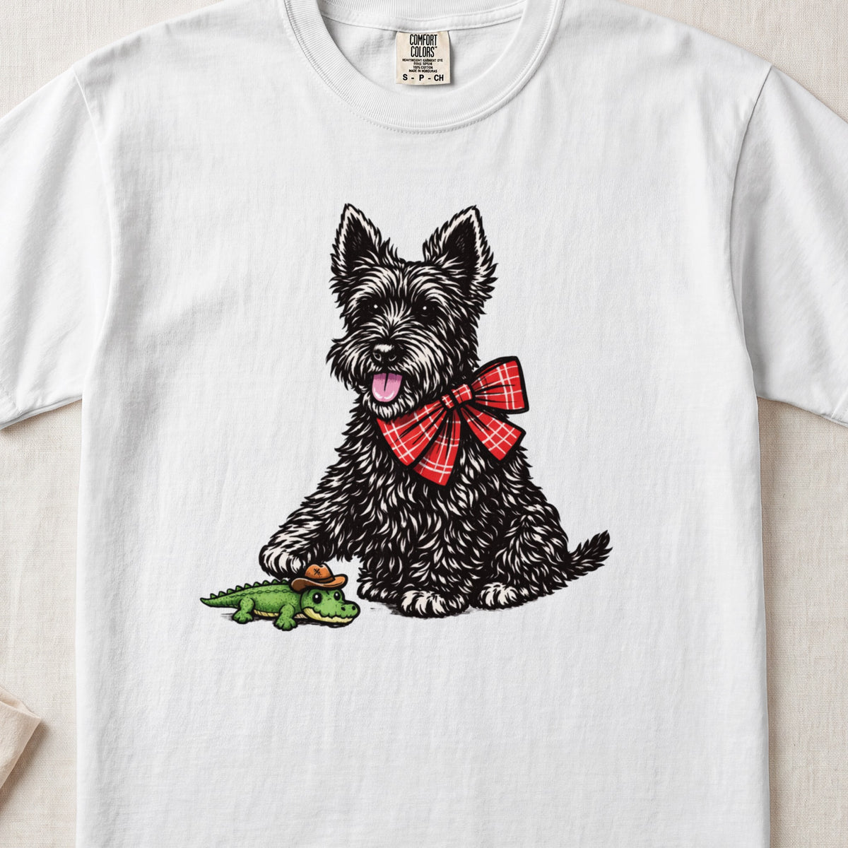 A white crewneck t-shirt featuring an illustration of a black Scottish Terrier wearing a red plaid bow, touching a small green alligator toy wearing a cowboy hat.
